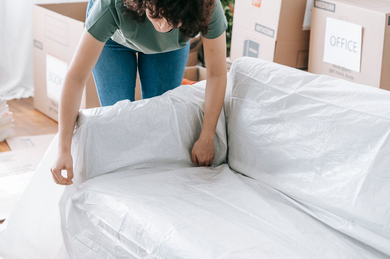 Une femme enveloppe avec soin un canapé dans un matériau de protection tout en préparant des cartons dans une pièce lumineuse.